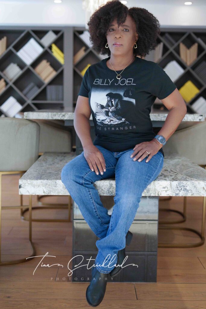 African American Woman sitting on a counter.  She's wearing a black graphic t-shirt, blue jeans, and black boots.